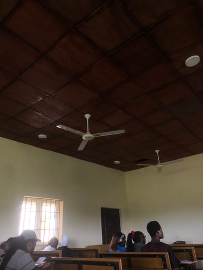 This media file captures an interior scene, likely a classroom or lecture hall, in Farin Yashe, Nigeria. The perspective is looking upwards towards a dark wood-paneled ceiling with a diamond-shaped pattern. Two white ceiling fans are visible, one closer and larger, with three blades, and another smaller one further away. A circular white light fixture is also present on the ceiling.
The lower portion of the image shows several people seated at desks, facing towards a bright window. The people are primarily young adults, with some appearing to be students. One individual in the foreground wears a dark baseball cap and a white shirt. Another person is partially visible behind them, also wearing a cap. Further back, a group of three individuals are more clearly visible. The person in the middle has glasses and a red hair accessory. The person to their right wears a blue top. Another individual further to the left is looking upwards and to the side, wearing a dark and red plaid shirt.
The scene suggests an ongoing activity, likely a lecture or lesson, given the classroom setting and the individuals seated at desks, seemingly engaged with the front of the room, which is implied to be towards the window. The bright light from the window suggests it might be daytime. The overall atmosphere appears focused and attentive. There is no visible text in the image.