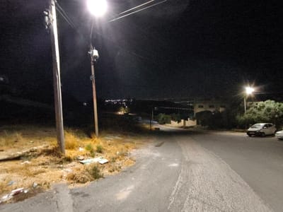 The image depicts a nighttime scene on a paved road in Umm Al-Quttayn, Jordan. Two wooden utility poles with lights are visible on the left side of the road. The pole in the foreground is closer and its light illuminates the immediate surroundings. The ground on the left is covered with dry grass and scattered debris, including what appears to be discarded packaging. Further down the road, a white car is parked on the right side, with another vehicle partially visible behind it. Buildings are visible in the background, illuminated by distant lights. The overall impression is of a semi-rural or suburban area at night.