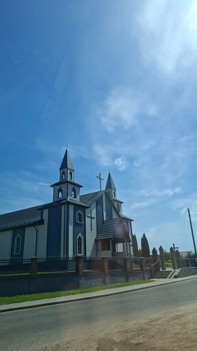 This is a photograph of the Church of the Assumption of the Blessed Virgin Mary in Dzyarzhynsk, Belarus. The church is a modern building with blue and white siding and a dark gray roof. It features two prominent towers, each topped with a cross, and a central entrance with a covered porch. A black metal fence with brick posts surrounds the property. The church is situated on a street with a paved road and sidewalk in the foreground. The sky is a clear blue with some wispy white clouds. Trees and other buildings can be seen in the background. The image appears to have been taken during the daytime on a sunny day.