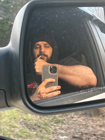 An image taken from outside a vehicle displays the reflection of a male individual in its passenger-side mirror. The scene is located in the Village of South Blooming Grove, United States of America.
The reflected individual is a male, appearing to be in his 20s or 30s, with a dark beard and mustache. He wears a dark hooded garment and a dark t-shirt, part of which shows a red and white graphic, including the letter "A". His right index finger is positioned at his upper lip. He is holding a smartphone, specifically an iPhone 13 Pro or 14 Pro model, in his left hand, angling it to capture the reflection. Tattoos are faintly visible on the knuckles of his left hand.
The mirror frame is light grey with black trim. Partially visible at the bottom right of the mirror's reflective surface is the etched warning "OBJECTS IN MIRROR ARE CLOSER THAN THEY APPEAR". The external environment behind and below the mirror shows a sparse, wooded area with bare trees and patches of green grass and dry vegetation on the ground.