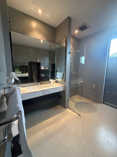 A modern bathroom features a glass-enclosed shower area adjacent to a vanity with a rectangular sink and dark cabinetry. A large mirror spans above the vanity, reflecting the bathroom's minimalist design. A chrome towel rack is attached near the sink. Natural light pours in through a window, illuminating the neutral-toned tile flooring and gray concrete walls.