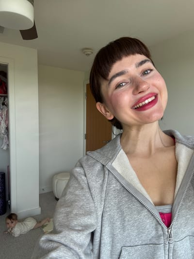 An indoor, vertical selfie captured from an overhead perspective, with the original image rotated 90 degrees counter-clockwise. The main subject is a woman with dark hair and bangs, a light complexion, and prominent eyebrows, smiling widely at the camera with red lipstick. Her head is tilted, and her green eyes are clearly visible. She is wearing a grey zippered hoodie, partially unzipped to reveal a bright pink garment underneath.
In the lower right background, an infant baby lies on its stomach on a light grey carpet, wearing a light-colored onesie and looking upwards. A small, patterned play mat is beneath the baby. The background primarily consists of a white wall and a light brown wooden door or wardrobe. A white smoke detector is mounted on the wall. A white dresser or cabinet is visible in the mid-ground, with some items (possibly clothes) stored within. The room appears brightly lit, suggesting it is daytime. No visible text is present. The overall mood conveyed by the woman's smile is cheerful.