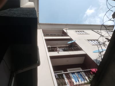 The image shows a multi-story building with balconies and windows, seen from a low angle. The building is light-colored, possibly cream or beige, with darker trim around the windows and balconies. There are several balconies visible, some with decorative metal railings. On the balconies, there are clothes hanging and what appear to be satellite dishes. Barbed wire is visible in the foreground, partially obscuring the view of the building. The sky above is blue with some scattered clouds. The scene appears to be in Alagbede, Nigeria, based on the context provided.