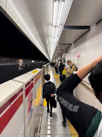 An angled view of a subway platform. Several people are waiting along the yellow safety line. Fluorescent lights illuminate the platform edge, running alongside a dark void. A person in the foreground is wearing a black shirt with white lettering on the sleeve. Red and white advertisements are visible on the right side of the platform. The scene is somewhat distorted due to the camera angle.