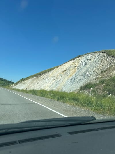 The image is a roadside view of a rocky embankment under a clear blue sky. The embankment is layered with rock of varying colors, including white, yellow, and gray. Green grass grows along the base of the embankment and on the top. A paved road with a white line marking the lane runs along the left side of the image. The foreground shows a portion of a car's dashboard and windshield wipers. The scene appears to be located in a natural, rural environment, likely a mountainous or hilly region. The location is specified as Aya, Russia.