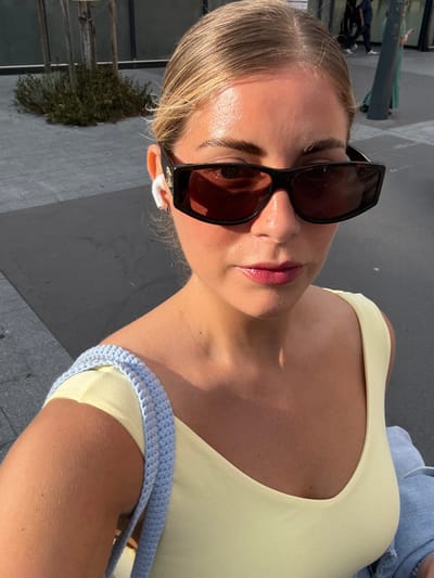 This is a close-up, sideways selfie of a young woman wearing sunglasses and a pale yellow tank top. Her blonde hair is pulled back, and she has light pink lipstick on. She is wearing small white earbuds. Her left shoulder is visible, and she has a light blue crocheted strap across her chest, which appears to be part of a bag. The background is a grey, paved surface, with some greenery and metal structures visible further back, suggesting an outdoor urban setting. The lighting suggests it is daytime, likely sunny, given the strong highlights on her skin and the sunglasses. The image appears to be taken in Saint-Ouen-sur-Seine, France.