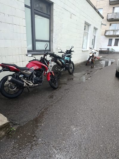 The image depicts a street scene in Khorol, Ukraine, with a focus on parked motorcycles and the surrounding environment. The foreground features a red and silver motorcycle parked on a wet asphalt surface. To the left, the wall of a white brick building is visible, with a large window. Behind the red motorcycle, a black and blue motorcycle is parked, with a license plate that reads "BI 8071 JA". Further down the street, a white dirt bike is parked next to a low concrete structure and a bench. In the background, parts of a multi-story building with balconies are visible. The ground is wet, indicating recent rain, with several puddles present. The overall impression is that of an outdoor, possibly residential or commercial, area in a Ukrainian town.