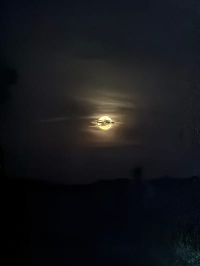 A blurry image captures a bright moon partially obscured by clouds, possibly taken through a window or wet surface. The background appears dark, with vague outlines of foliage visible at the bottom. A darker mass appears to the right, possibly a building or another large object. The location, city of Cotorra, Colombia, is not visibly determinable from the image.