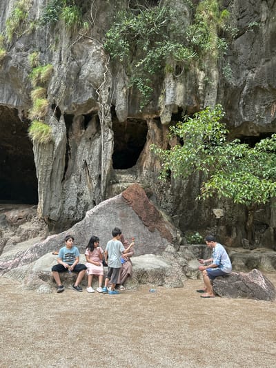 The image depicts a rocky cliff face with several cave openings in Tambon Krasom, Thailand. In the foreground, a group of four individuals, three children and an adult, are seated on a large rock. The two boys and the girl are sitting closely together, with the girl holding an ice cream cone. The adult, a man, is seated on a separate rock to the right, looking at his phone. A few bottles are scattered on the ground. The environment is natural, with vegetation growing on the rocks and around the cave entrances. The ground is covered with small stones and sand.