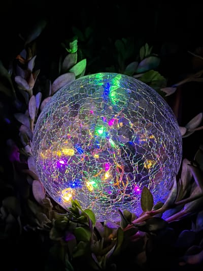 This is a close-up, low-angle photograph of a decorative crackle glass orb light illuminated by internal colorful LEDs. The orb is nestled amongst dark green and purple foliage. The glass has a network of fine cracks, which refract the light from within. The colors of the LEDs visible are green, blue, purple, pink, and yellow. The background is completely dark, suggesting the image was taken at night. The location is Uhornyky, Ukraine. There are no people or animals present in the image, and no discernible actions or interactions are occurring.