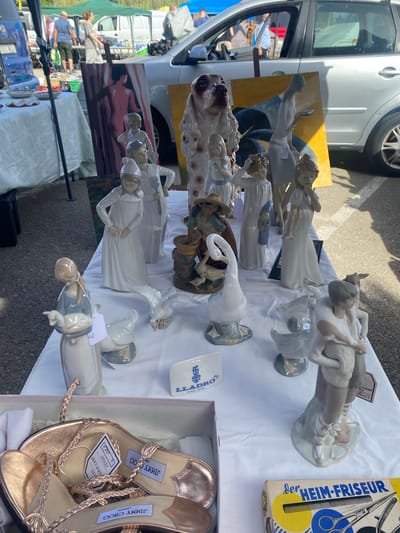 A market stall in San Luis de Sabinillas, Spain, displays a variety of ceramic figurines and a pair of rose gold sandals. The figurines, primarily in white porcelain, depict children, birds, and a large dog. One notable figurine is a woman holding a lamb, and another features a pair of dancing children. A painting of a nude figure is propped up behind the figurines. A box of hair salon tools, labeled "der HEIM-FRISEUR," is visible in the foreground. The stall is set up outdoors, with a white tablecloth covering the display surface. In the background, people can be seen browsing at other stalls, and vehicles are parked nearby. A tent canopy provides shade over part of the market.