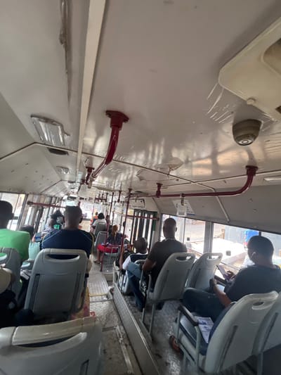 The image displays the interior of a public transit bus in Sumalia Town, Sierra Leone, observed from a forward-left perspective looking towards the rear. Approximately 10 to 15 adult passengers are visible, primarily seated, facing forward in light gray plastic seats. The bus ceiling is an off-white color, exhibiting areas of discoloration, possible water damage, and minor warping. Red curved metal handholds are mounted along the ceiling, which also features several rectangular light fixtures and a small, gray, dome-shaped security camera on the right side.
A person wearing a green shirt is seated in the left foreground. Directly ahead, a male in a dark blue shirt with yellow trim occupies a seat. In the mid-ground, a male in a black t-shirt on the right is engaged with a handheld device, with a white earbud visible. Another individual in a black shirt is seated in the mid-right. A woman in a red and white patterned top is visible in the mid-left. The bus floor features a metal, textured surface in the central aisle, where a single brown sandal is present on the right side. Large windows along the right side of the bus reveal an urban exterior with buildings and bright daylight.