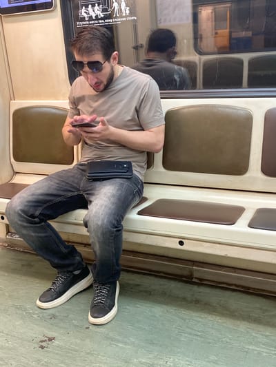 A man wearing sunglasses and a t-shirt sits on a subway seat in Gavrilkovo, Russia, looking at his phone. He has a black fanny pack across his waist. The subway car has brown seats and a green floor. A reflection of another person is visible in the window.