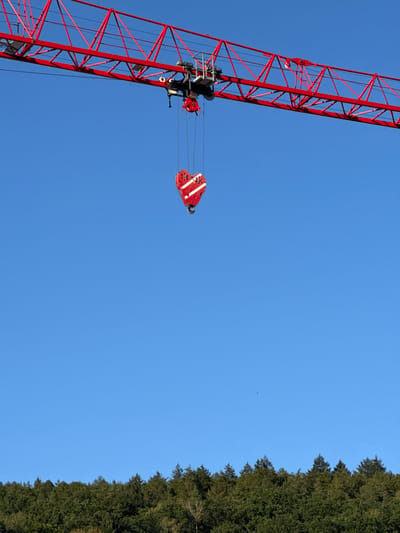 An image shows a red construction crane against a clear blue sky. The boom of the crane extends horizontally across the top portion of the frame, and from the trolley, a red heart-shaped object with white stripes is suspended by cables. Below the crane and the heart, a dense green forest stretches across the horizon. The image appears to be taken outdoors in daylight.