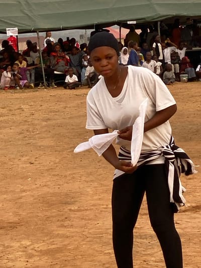 A person stands in a sandy area, wearing a white shirt, black leggings, and a black and white striped cloth around their waist. They are holding two white cloths. Behind them, a crowd of people are seated under a tent or awning, facing forward. The people in the crowd are diverse, with some wearing white and others wearing colorful clothing. The setting appears to be outdoors.