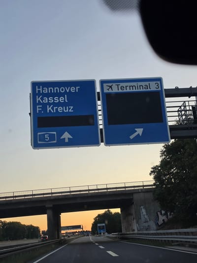 Two blue highway signs are visible in the foreground, mounted above a highway. The left sign indicates destinations: "Hannover", "Kassel", and "F. Kreuz" with the number "5" and an upward arrow. The right sign indicates "Terminal 3" with an airplane icon and an arrow pointing to the right. The signs are positioned over a multi-lane highway with traffic visible in the distance. An elevated bridge crosses over the highway. The sky is a pale orange, suggesting it is either dawn or dusk. The environment appears to be a highway interchange.