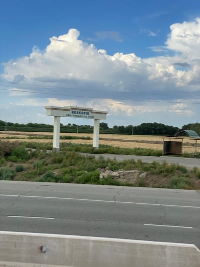 The image depicts a roadside scene in Leninizm, Uzbekistan. A paved road with white lane markings runs diagonally across the frame, disappearing into the distance. To the left of the road, a large, modern sign with two white pillars stands prominently. The sign bears text in a blue, sans-serif font: "BESKOPIR" is written in larger letters above "AWIL PUQARALAR JIYINI" in smaller letters. Beyond the sign, the landscape is characterized by fields of dry, golden vegetation, suggesting a rural or agricultural setting. A dark line of trees forms a backdrop to the fields, and a small, open-sided shelter with a curved roof is visible near the road. The sky is a vibrant blue, dotted with fluffy white clouds, indicative of daytime with fair weather. The image appears to be taken from a slightly elevated position, possibly from a moving vehicle, given the slight tilt of the frame. There are no people visible in the image. The overall impression is one of a quiet, open landscape in a rural part of Uzbekistan.