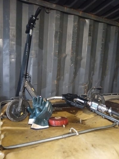 The image shows a black electric scooter leaning against a gray corrugated metal wall, likely inside a shipping container or a storage unit. A black fuel nozzle with a digital meter is lying on a tan plastic container, next to a pair of dark green gloves with white cuffs and a red plastic cap. A metal bar with a white rope tie is also present on the container. The location is assumed to be Яльгелево, Russia based on the prompt. The scene depicts unrelated items stored together in a utilitarian setting. No people are visible.