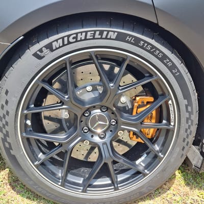 This media file is a close-up, eye-level shot of a car wheel, specifically a Michelin Pilot Sport tire mounted on a multi-spoke black AMG alloy wheel. The car appears to be dark gray. The wheel features bright orange brake calipers, also branded with AMG and "CARBON CERAMIC". The tire has prominent white lettering indicating its size "HL 315/35 ZR 21" and brand "MICHELIN". Additional text on the tire includes "MADE IN FRANCE", "EXTRA LOAD", "OUTSIDE", and various tire code markings. The wheel hub displays the Mercedes-Benz star logo. The car is parked outdoors on a grassy area, with some dried grass and sunlight visible on the ground and casting shadows on the wheel. The lighting suggests it is daytime, likely sunny. The setting appears to be a parking area or roadside given the presence of grass. No people are visible in the image. The focus is entirely on the automotive components, highlighting the tire, wheel, and braking system.