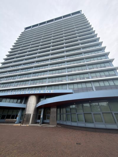 This media file captures a low-angle, wide shot of a modern, tall building in Rijswijk, Netherlands. The building features a distinctive facade with repeating vertical fins that create a sharp, angular silhouette against a bright, overcast sky. The facade is predominantly glass and metal, with rows of windows reflecting the sky and surrounding environment.
The building's name, "HOOGVOORDE," is prominently displayed vertically in large, white sans-serif letters on a blue accent panel on one side of the structure. Towards the base of the building, a brick-paved area is visible, with a curved, metallic structure integrating into the building's design. Signage in the lower portion indicates an address: "Bogardplein 15-37."
The scene appears to be set during the daytime, given the bright, diffused light. The weather seems to be cloudy or overcast. There are no people visible in the frame. The overall impression is one of modern architecture, with a focus on the building's geometric form and materials. The perspective emphasizes the height and imposing nature of the structure. The image is oriented such that the top of the building is to the left and the base is to the right.