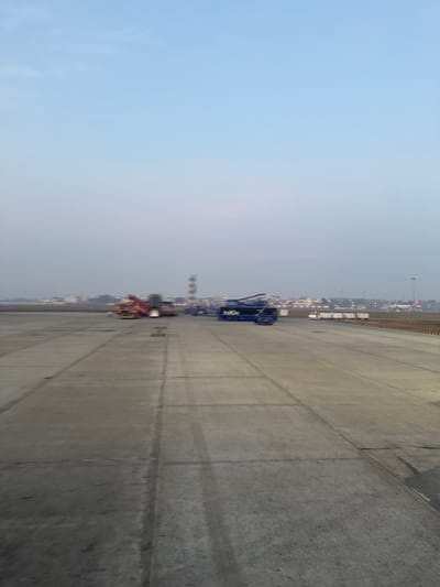 The image displays an airport tarmac, likely in daylight, with a clear blue sky dominating the left side. The tarmac is wide and gray, marked with expansion joints. Airport vehicles, including a blue vehicle with "IndiGo" branding, are parked alongside the tarmac. Some buildings and structures are visible on the horizon, suggesting an active airport environment. The perspective is from ground level, emphasizing the scale of the tarmac.