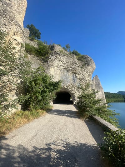 The image depicts a sunny day at a rocky cliff face with an opening that appears to be a tunnel or cave entrance. A paved path runs alongside the cliff. Green bushes and trees are growing on and around the rocks, and the path is lined with dry grass and shadows cast by foliage. In the distance, a body of water, likely a lake or river, is visible, bordered by lush green trees. The sky is a clear, vibrant blue. The scene suggests a natural, outdoor environment, possibly a hiking trail or a scenic viewpoint. There are no people or specific objects that indicate a particular activity, but the setting evokes a sense of exploration or peaceful contemplation. The time of day is clearly midday due to the strong sunlight and the sharp shadows. The location cues of the rocky terrain, water, and vegetation are consistent with a natural landscape, and the overall impression is one of a serene summer day. No text is visible in the image.