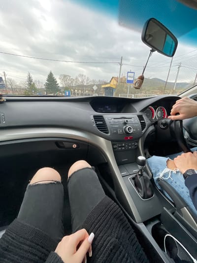 The image shows the interior of a car. A person is driving, and there's a passenger in the front seat. The dashboard and center console of the car are visible, featuring a stereo system and controls. The view through the windshield reveals a street scene in Susam, Bulgaria, with buildings, trees, and utility poles visible. The weather appears overcast.