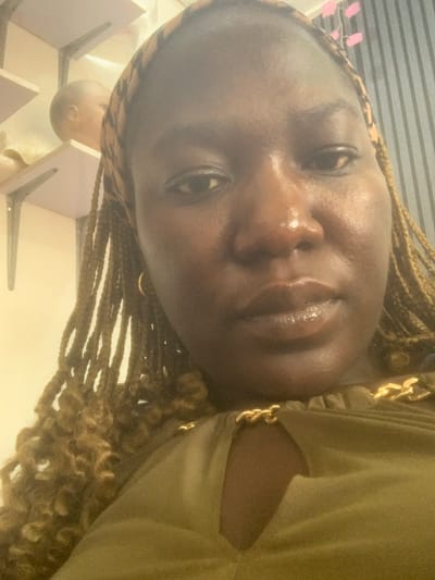 The image is a close-up selfie of a dark-skinned woman with braided hair. She is wearing a green top with gold chain accents. In the background, shelves are visible with a blurred object that appears to be a baby doll's head. The lighting is warm and casts a glow on the woman's face, highlighting her skin. The background is somewhat out of focus, drawing attention to the woman. The overall impression is of a casual, personal photograph taken indoors, possibly in a home environment in Rumuokoro, Nigeria.