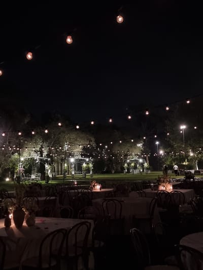 The image depicts an outdoor nighttime scene in Ga'ash, Israel, likely set up for an event. String lights with illuminated bulbs are strung across the dark sky, casting a warm glow. Below, a lawn area is dotted with tables and chairs, suggesting a dining or gathering space. Two ornate white gazebos, possibly part of the venue's architecture, are visible in the background, also illuminated by the string lights and ambient lighting. Several trees are scattered throughout the landscape, their foliage silhouetted against the night sky. A few people are present, including two individuals walking in the middle ground and others seated at tables in the foreground, though their faces are not discernible due to the low light conditions. The overall atmosphere is one of quiet festivity, with the lighting creating a magical ambiance.