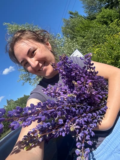 An outdoor, close-up, selfie-style photo shows a young woman with brown hair pulled up, smiling at the camera. She is holding a large bouquet of purple lupine flowers. The woman is wearing a dark blue t-shirt. Behind her, the background consists of lush green trees, a bright blue sky with a few white clouds, and a sliver of a gray car. A sign with Cyrillic text is visible in the background, slightly out of focus. The overall lighting suggests a sunny day. The setting appears to be a natural environment, possibly a roadside in Верх-Рождество, Russia.