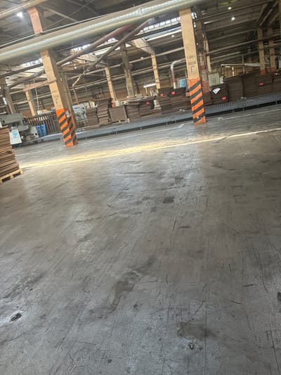 This is a wide, interior, eye-level shot of a large, industrial warehouse setting. In the foreground, the concrete floor shows scuff marks and debris, with a bright streak of sunlight illuminating the surface. In the mid-ground, stacks of cardboard boxes are visible, some on pallets, and others on a conveyor belt system. A piece of industrial machinery with a blue plastic component is also present. Structural elements like concrete pillars, some with orange and black hazard stripes and Cyrillic lettering, support the overhead pipes and ceiling. The background reveals more of the warehouse's industrial infrastructure, including more conveyor belts, lighting fixtures, and stacks of materials. The overall impression is that of a working factory or distribution center, possibly involved in cardboard production or packaging. It is not possible to determine the specific location in the city of Kashira, Russia from the image alone.