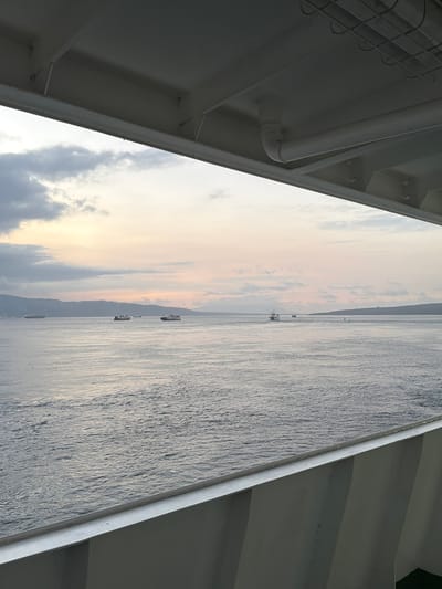 The image depicts a seascape viewed from the deck of a vessel. The perspective is angled, with the foreground dominated by the white structures of the ship, including pipes and beams. Beyond the ship's railings, a wide expanse of water stretches towards a distant, silhouetted landmass, likely a coastline. The water is textured with ripples, reflecting the soft, diffused light of dawn or dusk. Several small boats are scattered across the water, some appearing as dark silhouettes against the brighter sky, others with faint lights.
The sky transitions from a pale, luminous yellow-orange near the horizon to a soft blue with scattered, wispy clouds. The overall atmosphere is tranquil and serene, suggesting a peaceful moment at sea. The time of day is either early morning or late evening, given the low angle of the sun and the soft lighting. The location is identified as Melaya, Indonesia, implying a tropical or subtropical setting. No people are clearly visible in the image. The scene evokes a sense of calm and openness, with the vastness of the ocean and sky dominating the view. There is no visible text in the image.