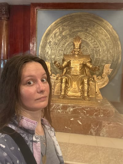 The photograph, taken indoors, features a woman standing in the foreground with a golden statue of a seated figure behind her. The statue is imposing, with intricate details on its armor and a large, circular, ornate background. The woman is facing the camera with a surprised expression, her head slightly tilted. She has long, dark brown hair and is wearing a patterned jacket over a gray shirt, with a necklace visible. The background shows elements of what appears to be a museum or a temple, with wooden paneling on the left and a marbled base for the statue. The specific location in the city of Hy Cương, Vietnam, is not evident from the image itself, but the style of the statue suggests a historical or cultural setting within Vietnam.