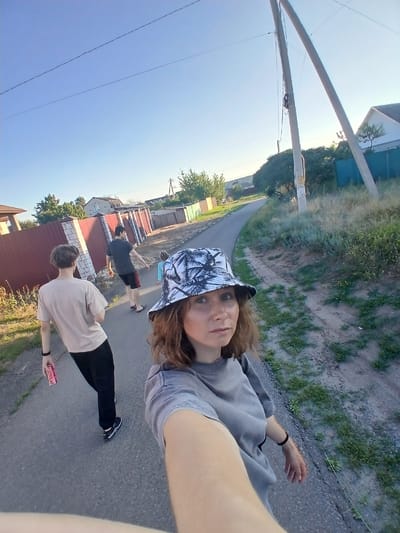 The image shows a selfie taken by a woman wearing a bucket hat, in what appears to be a rural or suburban area. She is walking on a paved road, and behind her, two males and a female are also walking on the road. One of the males is holding a red can. The road is lined with fences and houses. The sky is clear and blue, and the sun is shining, indicating it is daytime. The general setting suggests a residential area possibly in a village or small town, consistent with locations like Maslova Pristan', Russia, which is known for its rural landscapes and residential communities.