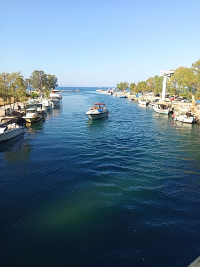 This is a wide-angle, eye-level outdoor shot of a clear blue sky above a channel of calm, dark blue water, likely in Georgioupoli, Greece, given the context. The channel is lined on both sides with moored boats and trees. On the left bank, there are several white and grey motorboats of various sizes, some appearing to be fishing vessels and others more recreational. A paved walkway runs along the bank, with streetlights and some parked cars visible behind the boats. On the right bank, there is a similar arrangement of moored boats, including some larger white vessels. A tall, white crane structure is prominent on this side, and more parked cars are visible behind the boats and trees. A few people are visible on the boats, one group lounging on the stern of a moving boat in the middle of the channel. The water is mostly a deep blue, with lighter turquoise hues near the edges and where sunlight penetrates. The distant horizon shows the sea meeting the sky, with two small lighthouses visible on either side of the channel entrance. The overall atmosphere is peaceful and sunny.