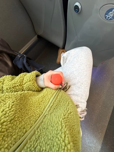The image depicts a seated individual within a public transport vehicle, potentially operating in or around Crumlin, United Kingdom, though no specific external landmarks are visible to confirm the exact location. The subject is partially visible from the upper chest to the knees, wearing a light green, textured long-sleeved jacket over a gray long-sleeved top and light gray sweatpants. The right hand, featuring decorated fingernails, holds a small, red cylindrical container with a flat top. A fringed, light-colored scarf is partially visible beneath the right arm. To the individual's left, a dark brown shoulder bag contains a folded dark blue garment. The vehicle interior features gray upholstered seating and gray wall panels. One panel includes a circular silver USB charging port with both Type A and Type C inputs, and a circular emblem depicting a stylized figure in a wheelchair. The floor is dark gray. The individual's right foot, clad in a brown flat shoe, rests on the floor.