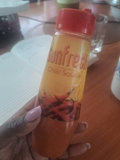 A dark-skinned hand with manicured, light purple, glittered fingernails is holding a clear plastic bottle of "Sunfresh Chilli Sauce." The bottle features a red screw-top cap and contains a reddish-brown granular substance and an illustration of red chili peppers. The hand is positioned centrally, gripping the bottle with the thumb and fingers. The scene is set indoors on a polished wooden surface, likely a table. In the blurred background, an open white notebook with lined pages is visible on the left, and a clear drinking glass along with other indistinct objects are present on the right. The location is Muthambi ward, Kenya.