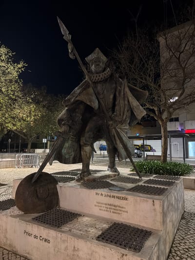 The image presents a bronze statue of António, Prior of Crato, located in Largo do Prior do Crato, Santarém, Portugal, at night. The statue depicts a male figure standing upright, clad in historical attire consisting of a ruff collar, a layered cape, and a hat. The figure holds a long spear vertically, with its pointed head extending upwards beyond the frame and its base resting on a large, spherical bronze object positioned at the front left of the statue's base.
The statue is mounted on a multi-tiered, light-colored concrete pedestal. The lower front of the pedestal is inscribed with "Prior do Crato." The upper front face displays the date "XIV de Julho de MDLXXX" (July 14, 1580), followed by a Portuguese inscription: "Sejamos hoje todos aqui doados seja quem o seja que todos seremos viva el-Rei D. António I." The top surface of the pedestal features numerous rectangular, dark-colored, textured metal plates arranged in a grid pattern.
The surroundings are illuminated by artificial light sources, casting the dark night sky as the backdrop. The ground is paved with cobblestones. To the left of the statue, several metal crowd barriers are visible, beyond which are streetlights and trees with green foliage. To the right, a modern building with illuminated windows (displaying red and blue light accents) is present, featuring a "GARAGEM" sign and a small, bare-branched tree in front of it. Low green foliage is planted at the base of the building. Distant vehicles are faintly visible on the street in the background. No other individuals are present within the immediate scene.