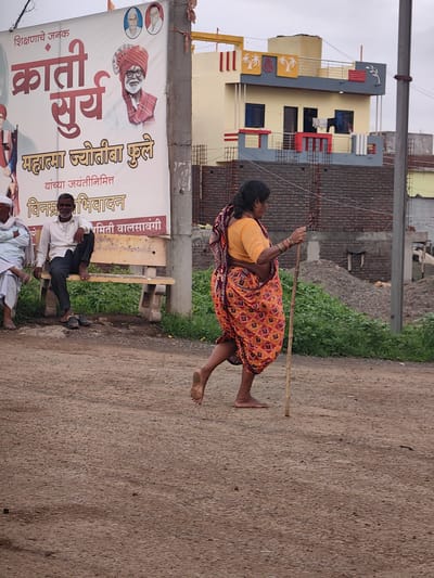 In Walsawangi, India, a woman in a vibrant orange and red patterned sari walks with a wooden staff. She is barefoot and appears to be moving from right to left across a dirt road. Behind her, a large banner with Marathi text and portraits of men is displayed on a pillar. To the left of the banner, two men are seated on a concrete bench. One man wears a white shirt and dark pants, while the other is dressed in a white garment. In the background, a multi-story building with balconies and decorative elements stands against an overcast sky, with piles of gravel and greenery visible.