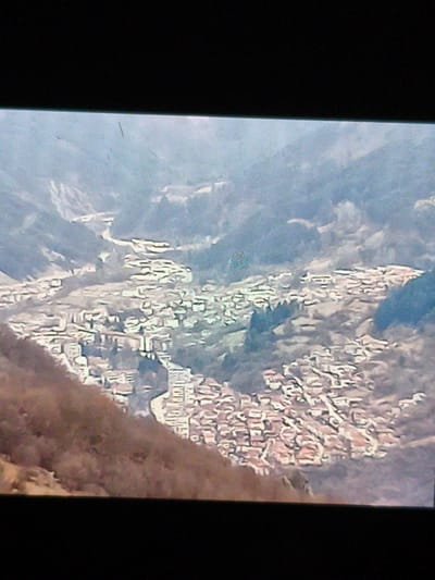 A high-angle shot shows the city of Devin, Bulgaria. The city lies in a valley surrounded by mountains. The roofs of buildings in the town appear red. A river runs through the center of the town. A brown, barren hillside occupies the lower-left corner of the image. The overall atmosphere is hazy.