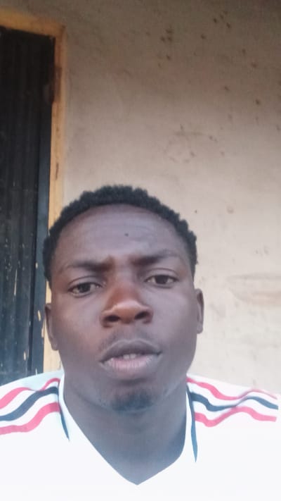 A young Black male with short, dark hair is captured in a close-up, eye-level selfie. He is wearing a white t-shirt with red, white, and blue striped detailing on the shoulders. The background appears to be an exterior wall, possibly made of concrete or stucco, with some subtle discoloration and small dark spots. To the left of the frame, a dark-colored door with vertical slats is visible, framed by a light-colored wooden trim. The lighting suggests it is daytime, with natural light illuminating the scene. The image is taken outdoors, likely in an urban or semi-urban environment within the city of Donga, Nigeria.