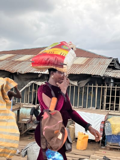 In Oko Amakom, Nigeria, a woman with dark skin wears a dark maroon velvet dress with cutouts on the sleeves. She carries a large bag of rice on her head and a brown backpack with pink accents over her shoulder. Her right hand is near her face, possibly touching her nose or adjusting her mask. In the background, a dilapidated building with a rusted corrugated metal roof is visible. A man sits on a wooden structure in front of the building, his legs bare. Another person, mostly obscured, wears a yellow and orange striped garment on the left side of the frame. The ground is made of wooden planks, and various objects, including an orange plastic container and a grey tarp, are scattered around. The sky is overcast with clouds.