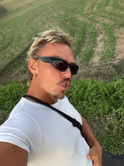 A selfie taken outdoors in daylight features a fair-skinned individual with blonde hair, wearing sunglasses and a white t-shirt with a black strap across the chest. The background consists of green grass and vegetation. The location cannot be determined to be within Selemadeg Timur, Indonesia, based on the provided image.
