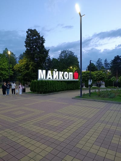 A group of five people, including two adults and three children, are walking on a paved pathway in a park-like setting. The pathway is made of interlocking square pavers in shades of yellow and dark red. In the background, a large, illuminated sign reads "МАЙКОП" (Maykop) in white block letters, with a red apple-shaped object serving as the last letter. The sign is set against a backdrop of manicured green hedges. A tall street lamp with a bright light stands to the right of the sign. Trees with lush green foliage are visible throughout the scene, and the sky above is a dusky blue with scattered clouds. The overall atmosphere suggests an evening setting in a public recreational area. The location is identified as the city of Maykop, Russia.