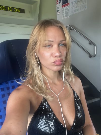 This media file is a portrait of a woman with blonde hair and blue eyes, taken inside a train. She is wearing a black sequined top and white earphones. The woman is looking directly at the camera with a neutral expression. The background shows the interior of a train carriage, with a metal handrail visible on the left, and a blue patterned seat. A digital display on the left side of the frame shows the time "16:36". There is also a poster with maps and text in German on the wall. The lighting suggests it is daytime. The location is identified as Bockhorn, Germany.