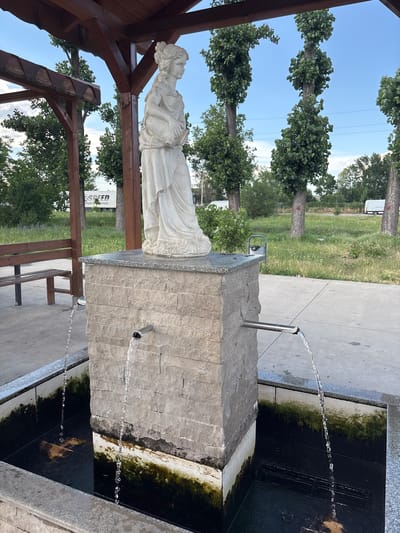 The image shows a stone fountain with a statue of a woman, possibly religious, in a park-like setting. Water flows from spigots into a dark basin that appears to have some algae growth. The statue is positioned on a wooden pergola structure, looking to the left. In the background, there are tall trees with green foliage, a paved walkway, and a grassy area. A few white vans are visible in the distance. The sky is blue with some clouds, suggesting it is daytime. A bench is visible in the lower right portion of the image, and another wooden structure is to the left of the fountain. A sign with text is partially visible behind the statue, reading "SPEED SPORT TRADITION." The overall scene suggests a public space, possibly a park or a rest stop. The time of day appears to be late afternoon due to the soft lighting. The weather seems pleasant. The location context indicates Kazichene, Bulgaria.