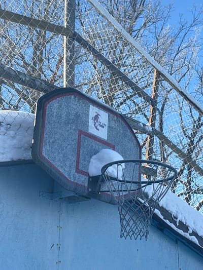 An outdoor basketball hoop and backboard are depicted under clear blue skies in a winter setting in Toksovo, Russia. The light gray, rectangular backboard features a red outline and a logo in the upper center with text "R-u-m" and a graphic of a figure on skis or a snowboard. A significant accumulation of white snow rests on the upper portion of the backboard, partially obscuring the attachment point of the black metal hoop. The hoop is fitted with a white net, appearing stiff. The backboard is mounted on a light blue-grey wall. In the background, a chain-link fence structure, partially covered in snow, spans the upper frame, behind which bare deciduous trees are visible against the bright blue sky. Additional snow covers a horizontal surface to the immediate left of the backboard and along elements of the fence structure. No individuals are present.