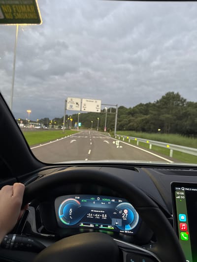The image depicts a first-person view from inside a vehicle, specifically from the driver's perspective, on an access road leading to an airport. The location is identified as Santander Airport (SDR) in Camargo, Spain, based on the prominent "Terminal" signage typical of airport approach roads.
The scene is set in the late evening, indicated by the car's digital dashboard display showing "22:06." The sky above is overcast.
**Objects and Subjects:**
- **Vehicle Interior:** A human left hand is positioned on the black, leather-wrapped steering wheel, indicating a driver. The digital instrument cluster displays a speed of "27 km/h," the vehicle in "D" (Drive) gear, "HEV" (Hybrid Electric Vehicle) mode, and an odometer reading of "8613.4 km." A head-up display shows "27" km/h on the windshield. To the right, an infotainment screen shows "10:06," a "5G" cellular signal, and applications including FaceTime, Maps, Music, and Phone. A yellow "NO FUMAR NO SMOKING" sign is partially visible through the upper left windshield.
- **Roadway:** The vehicle is on a paved, multi-lane road with clear white lane markings and directional arrows indicating straight ahead. The road features reflective road markers along the median and yellow reflective markers on the guardrail along the right shoulder.
- **Signs:** A large overhead road sign indicates directions to "Terminal," "Parking," "Bus," and "Taxi."
- **Environment:** Streetlights illuminate the road ahead and the distant airport infrastructure on the left. A grassy median separates the road, and a dense line of trees borders the right side of the roadway. Lights from what appear to be terminal buildings or parking structures are visible in the distance on the left.
**Actions and Interactions:**
The presence of a hand on the steering wheel indicates the driver is actively operating the vehicle, presumably driving towards the airport terminal. The displayed speed of 27 km/h suggests slow movement or a stop.