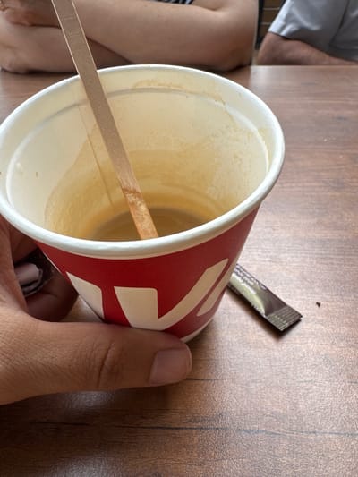 An individual's hand holds a red and white paper coffee cup containing a small amount of coffee and a wooden stirrer. The cup is positioned on a wooden table. Next to the cup is a silver packet of sugar. In the background, two individuals are seated at the table. The scene appears to be an indoor dining area.