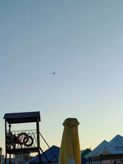 The image captures a view of the shoreline at Toranghylyk, Kazakhstan, under a pale blue sky. In the foreground, a lifeguard tower stands to the left, its dark roof and railing providing a stark contrast against the sky. Two life preservers are visible on the tower's platform. To the right of the tower, a folded yellow umbrella dominates the mid-ground, its fabric draped in a conical shape. Further to the right, a row of peaked tents or awnings extends into the distance, with some displaying text. A single bird is silhouetted in flight against the sky in the upper center of the image. The lower portion of the image shows a glimpse of distant buildings and foliage.